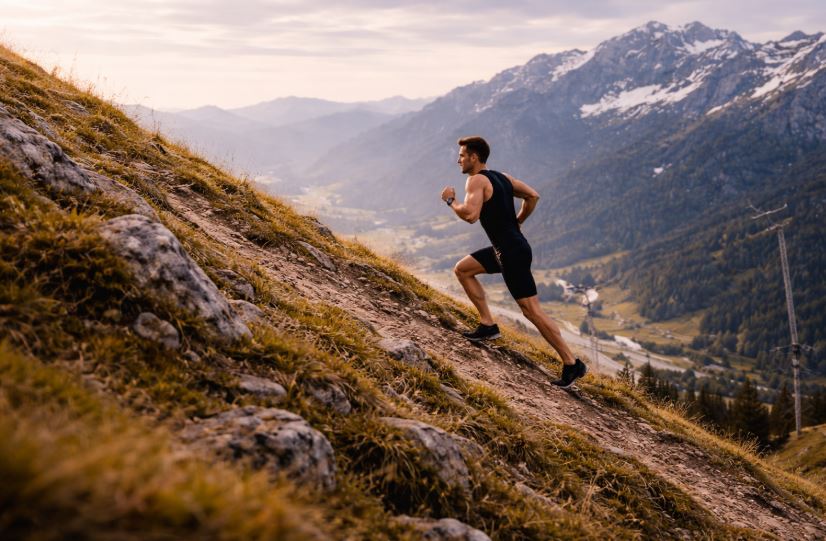 Lauftraining am Berg: Mann läuft von links nach rechts einen steilen Berghang hinauf, schwarzes Laufoutfit, Tal mit Wiesen, Fluss und Alm im Hintergrund, schneebedeckte Berge und Seilbahn in der Ferne